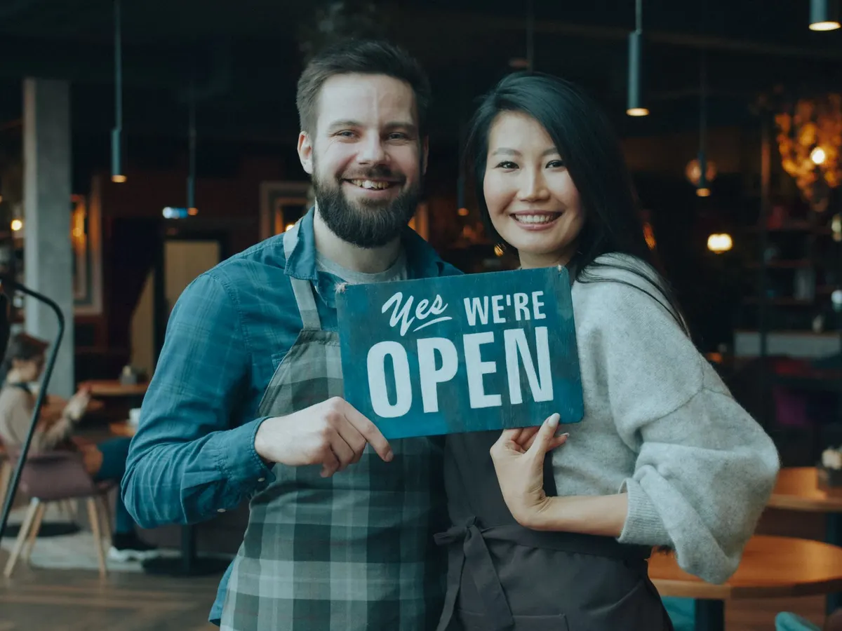 Ein Mann und eine Frau stehen lächelnd in ihrem Laden und halten ein Schild mit der Aufschrift „Yes, we’re open“.