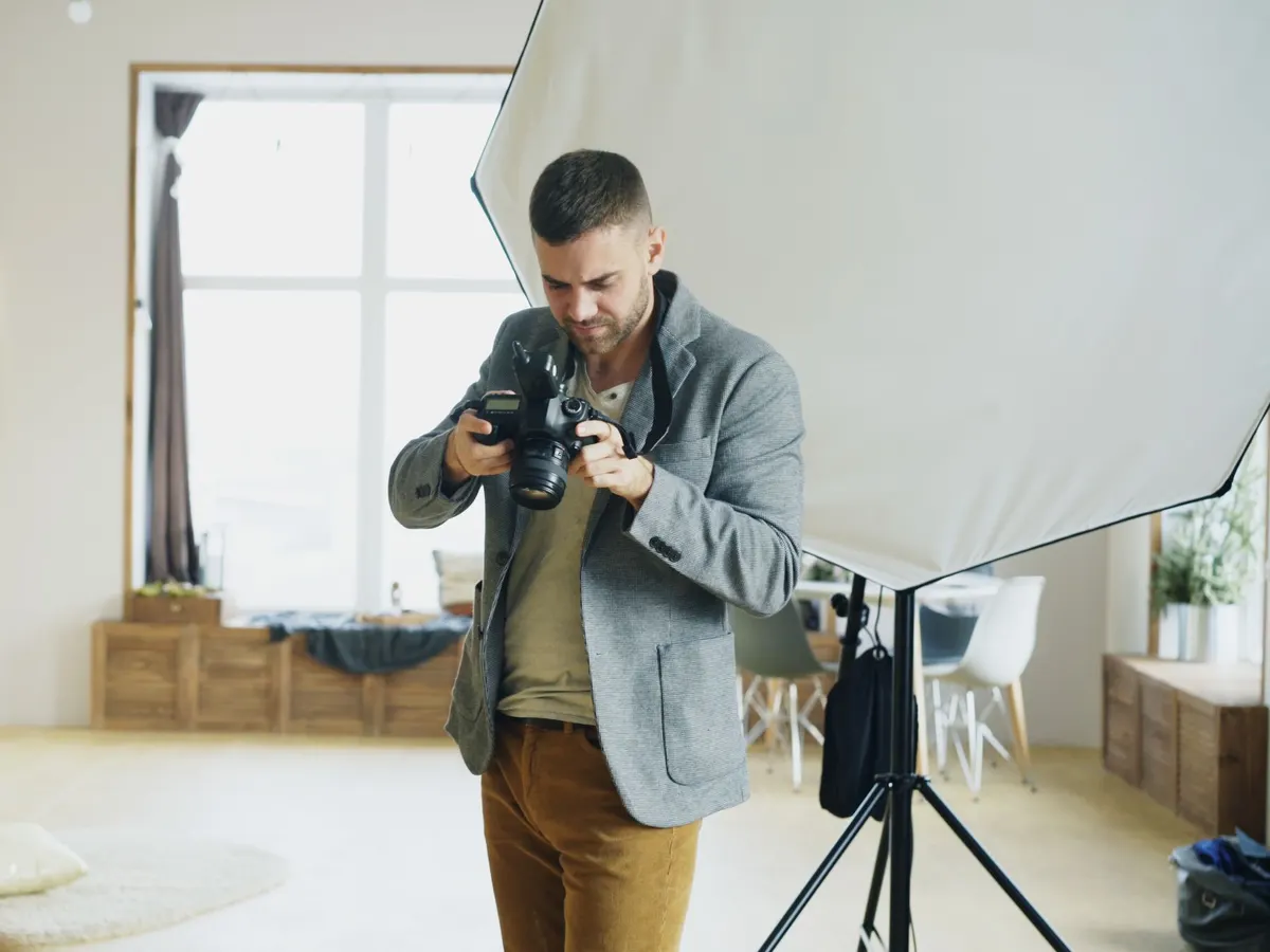 Ein Fotograf schaut im hellen Studio auf das Display seiner Kamera, neben ihm ein Stativ und eine Softbox.