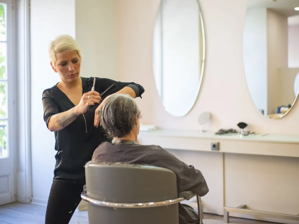 Friseurin schneidet einer älteren Kundin in einem hellen Salon mit runden Spiegeln die grauen Haare.