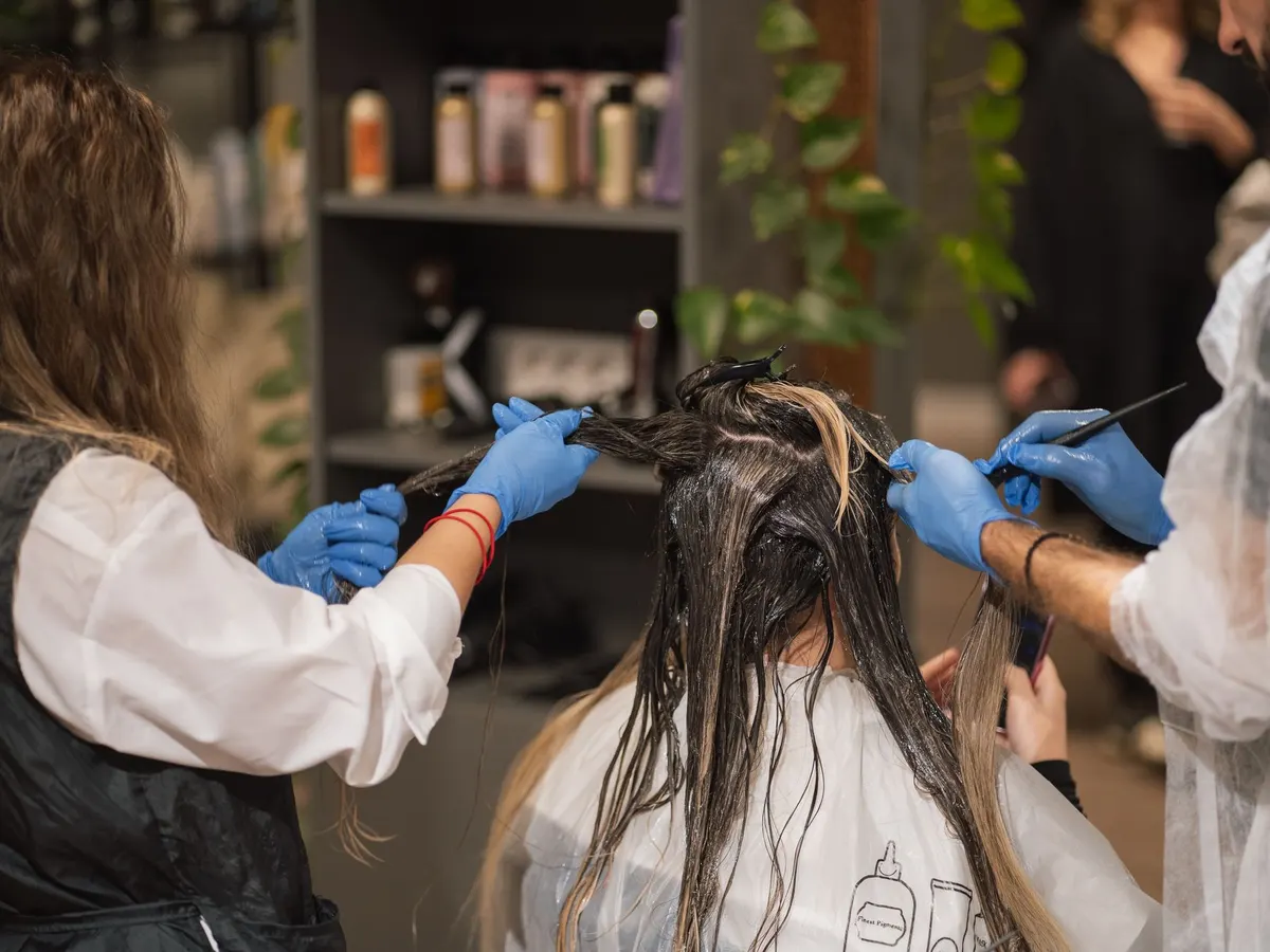 Zwei Friseurinnen mit Handschuhen tragen einer Kundin mit langen Haaren Farbe auf.