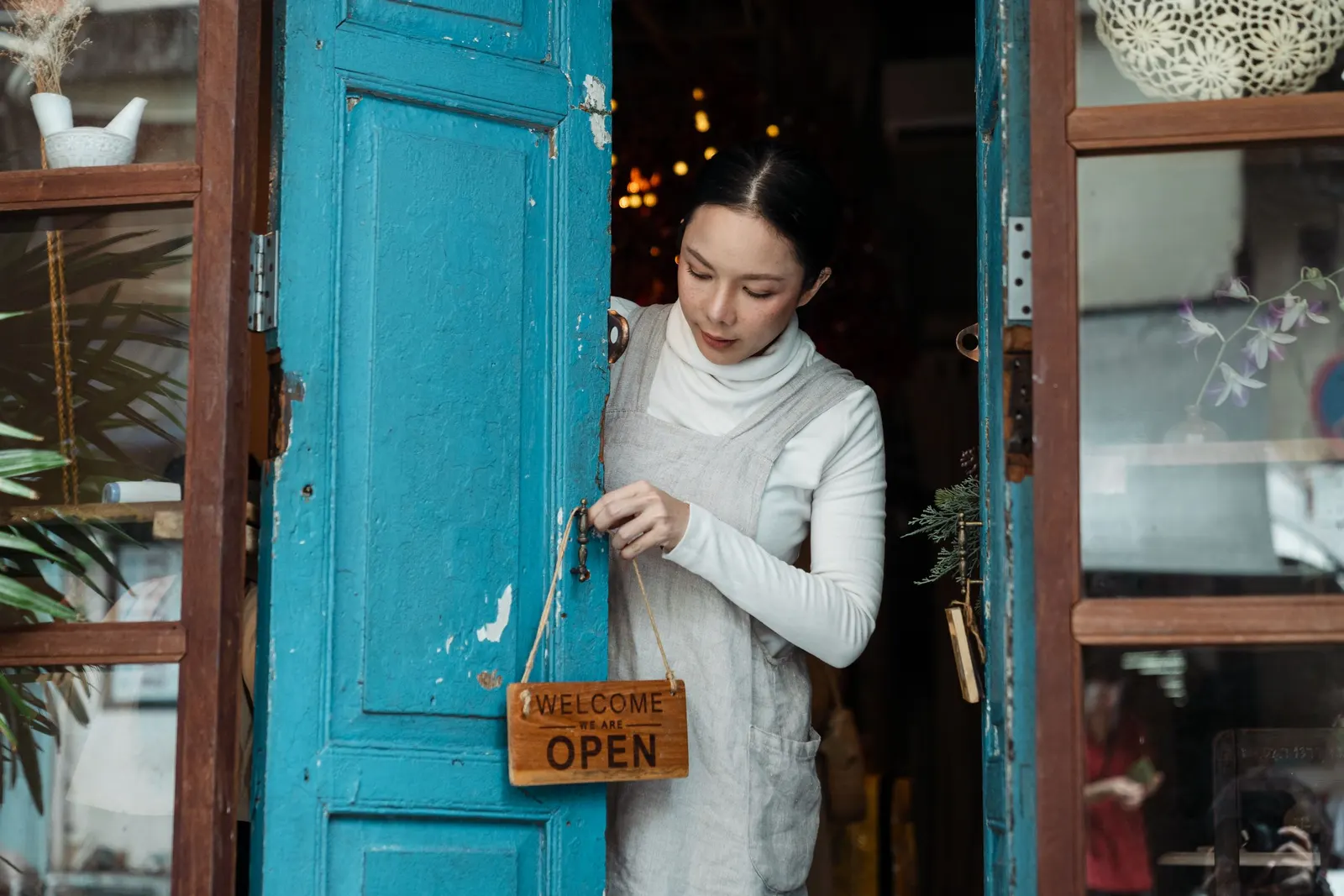 Eine Frau in Schürze öffnet die blaue Holztür ihres Cafés und hängt ein Schild mit der Aufschrift Open an den Türgriff.