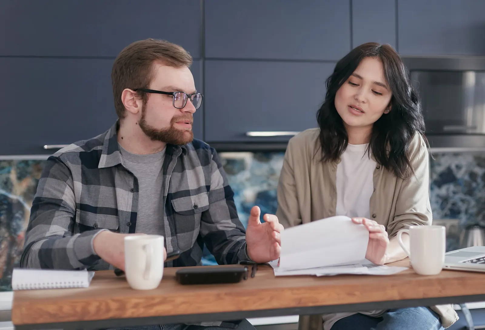 Ein Mann und eine Frau sitzen mit Kaffeetassen, Unterlagen und Taschenrechner am Küchentisch und besprechen ein Schreiben.