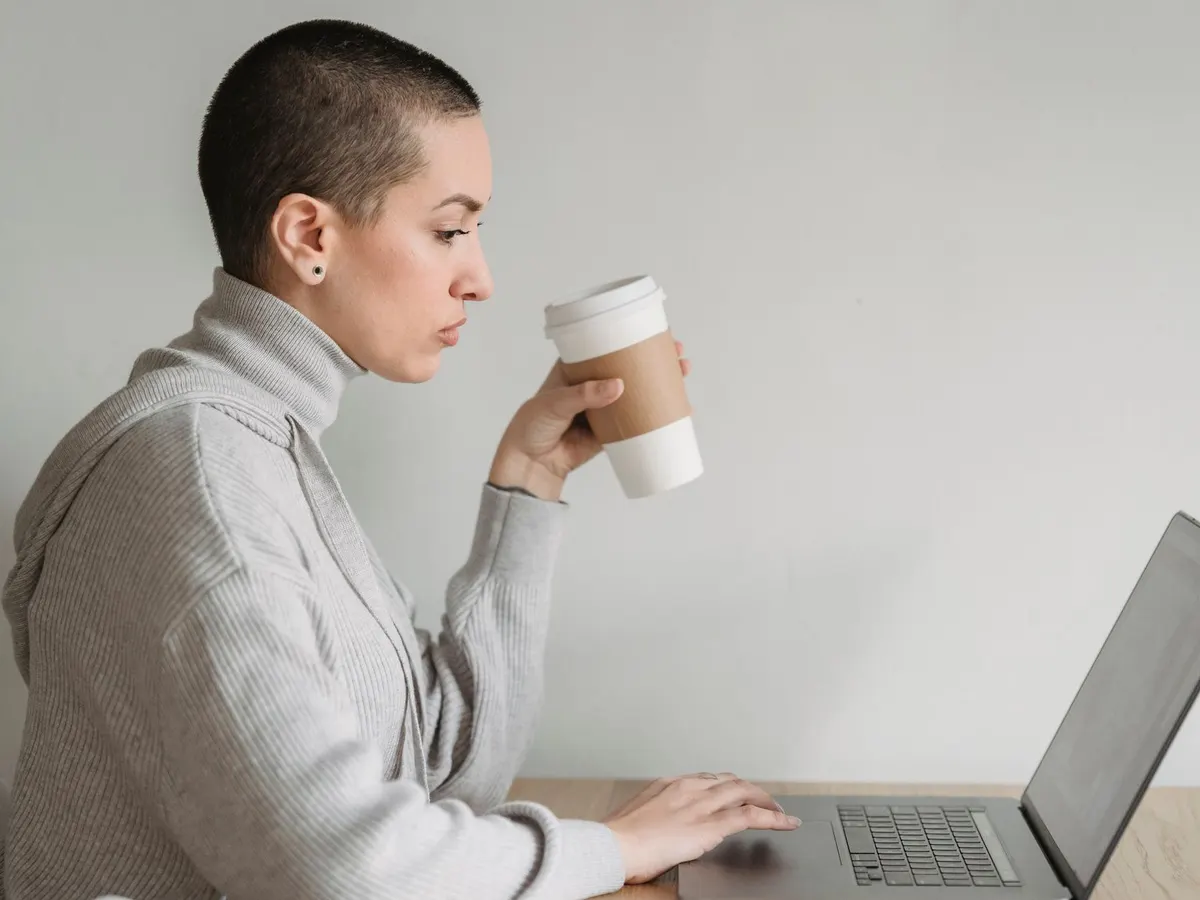 Eine Frau im grauen Pullover trinkt einen Kaffee und arbeitet dabei entspannt an ihrem Laptop.