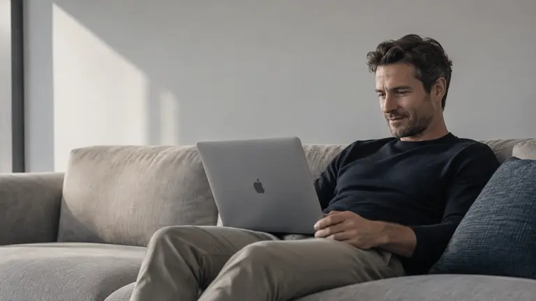 Mann sitzt auf Couch und blickt lächelnd auf MacBook auf seinem Schoß
