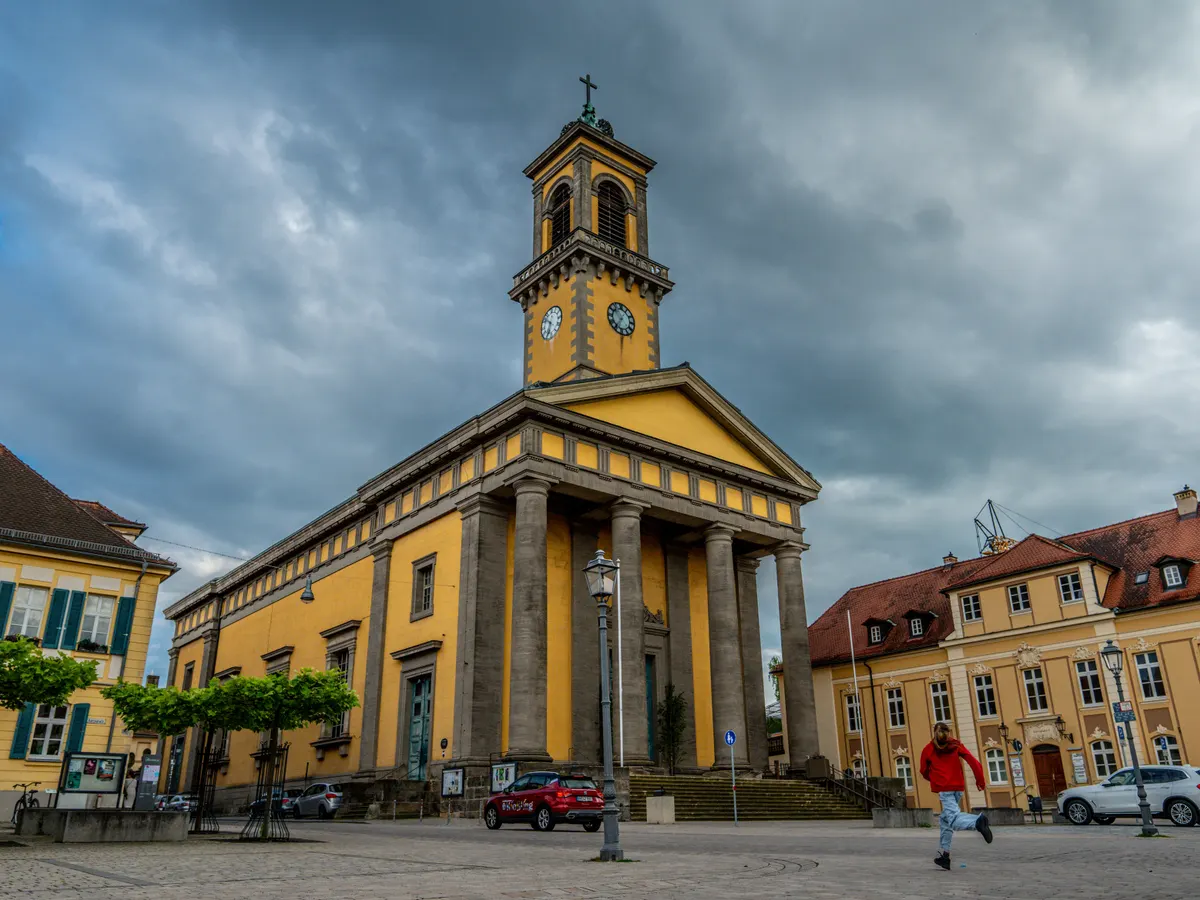 Die gelbe Kirche St. Ludwig in Ansbach mit Säulenvorbau und Glockenturm unter einem bewölkten Himmel, davor ein gepflasterter Platz.