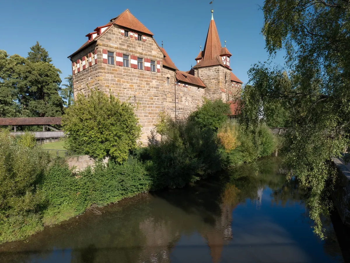 Das Wenzelschloss in Lauf mit Sandsteinmauern und spitzem Turm über der Pegnitz, die Bäume am Ufer spiegeln sich im ruhigen Wasser.